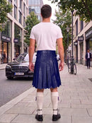 A man standing on a city sidewalk, viewed from behind, wearing a white t-shirt, a dark blue pleated leather kilt, white knee-high socks, and black shoes.
