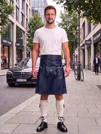 A man stands on a city sidewalk wearing a dark navy blue leather kilt with a black leather belt and silver buckle, paired with a white t-shirt, white knee-high socks, and black leather shoes.