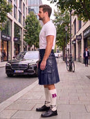 A man standing on a city sidewalk wearing a dark navy blue leather kilt, a white t-shirt, white socks with a pink accent, and black leather shoes. A black Mercedes-Benz car is parked on the street behind him.