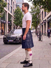 A man standing on a city sidewalk wearing a dark navy blue leather kilt, a white t-shirt, white socks with a pink accent, and black leather shoes. A black Mercedes-Benz car is parked on the street behind him.