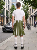 A man stands on a city sidewalk facing away from the camera, wearing an olive green pleated kilt with white vertical stripe panels, a white t-shirt, white knee-high socks, and black shoes. Storefronts, trees, and parked cars are visible in the background.