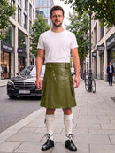 A man stands on a city sidewalk wearing a white crew-neck t-shirt and an olive green pleated leather kilt with silver hardware, paired with white knee-high socks and black leather shoes.