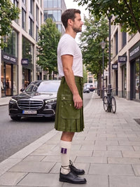 A man standing on a city sidewalk wearing a brown pleated kilt, white t-shirt, white socks, and black leather oxford shoes, with a street scene and parked cars in the background.