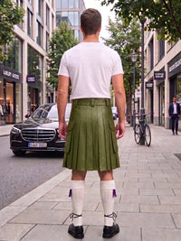 A man standing on a city sidewalk, viewed from behind, wearing an olive green pleated kilt with a white t-shirt, white knee-high socks, and black shoes. Buildings, trees, and parked cars are visible in the background.