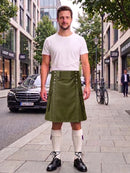 A man stands on a city sidewalk wearing a white crew-neck t-shirt and an olive green corduroy kilt with white knee-high socks and black leather shoes. The background shows a street scene with a black Mercedes-Benz, trees, and storefronts.