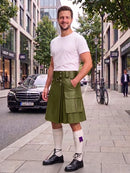 A man stands on a city sidewalk wearing a white crew-neck t-shirt, an olive green faux-leather kilt with a buttoned waistband, white knee-high socks, and black leather lace-up shoes. The background shows a blurred street scene with a Mercedes-Benz car, storefronts, and trees.