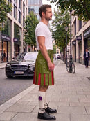 A man standing on a city sidewalk wearing a green kilt with red pleats, a white t-shirt, white socks, and black leather shoes, with a street scene in the background.
