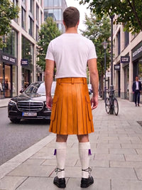 A man standing on a city street, viewed from behind, wearing a white t-shirt and a bright orange pleated leather kilt with white knee-high socks and black shoes.