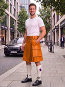 A man standing on a city sidewalk wearing a bright orange-brown leather kilt with a white t-shirt, white knee-high socks, and black leather shoes. A Mercedes-Benz car and city buildings are visible in the background.