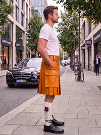 A man standing on a city sidewalk wearing a white t-shirt, a mustard yellow leather kilt with front pockets, white socks, and black leather shoes. A black Mercedes-Benz car is parked on the street behind him, with trees and storefronts in the background.