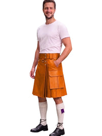 A man modeling a tan leather kilt with a matching belt, paired with a white t-shirt, white knee-high socks, and black shoes, on a solid white background.