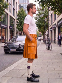 A man standing on a city sidewalk wearing a tan leather kilt, white t-shirt, white knee-high socks, and black leather shoes. The background shows a street with a black Mercedes-Benz car, trees, and buildings.