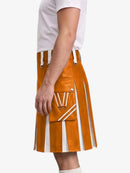A side view of a model wearing a mustard orange faux-leather kilt with white contrast stitching, pleats, and a front utility pocket, paired with a white t-shirt.