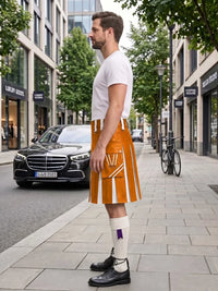 A man standing on a city sidewalk wearing a bright orange and white pleated kilt, a white t-shirt, white knee-high socks, and black leather shoes. A black Mercedes-Benz car is parked on the street behind him, with trees and storefronts in the background.