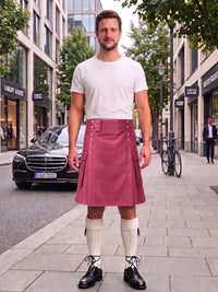 A man stands on a city sidewalk wearing a white crew-neck t-shirt and a dusty rose pleated kilt with silver metal accents. He is also wearing white knee-high socks and black leather shoes.