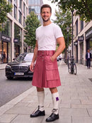 A man stands on a city sidewalk wearing a pink leather kilt with a matching belt, a white t-shirt, white knee-high socks, and black shoes. Buildings, trees, a parked black car, and a bicycle are visible in the background.