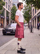A man standing on a city sidewalk wearing a pink pleated kilt, white t-shirt, white socks, and black leather shoes, with buildings, trees, and a parked black car in the background.