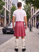 A man standing on a city sidewalk, viewed from behind, wearing a white t-shirt and a pink pleated leather kilt with white knee-high socks and black shoes.