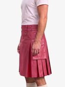 A man wearing a pink faux-leather kilt with pleats, flap pockets, and metal hardware, paired with a white t-shirt and white socks, shown from the waist down against a light gray background.