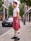 A man standing on a city sidewalk wearing a pink pleated kilt, white t-shirt, white knee-high socks, and black shoes, with buildings, trees, and a parked black car in the background.