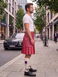A man standing on a city sidewalk wearing a pink pleated kilt, white t-shirt, white socks, and black shoes, with storefronts, trees, and parked cars in the background.