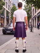 A man standing on a city sidewalk, viewed from behind, wearing a white t-shirt, a pleated purple kilt, white knee-high socks, and black shoes. Buildings, trees, a black Mercedes, and a bicycle are visible in the background.