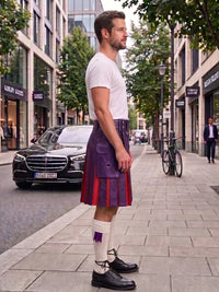 A man standing on a city sidewalk wearing a purple and red pleated kilt, a white t-shirt, white socks, and black leather shoes, with buildings, trees, and parked cars in the background.
