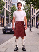 A man stands on a city sidewalk wearing a white short-sleeve t-shirt and a dark reddish-brown leather kilt, paired with white knee-high socks and black leather shoes. A black Mercedes-Benz car and storefronts are visible in the background.