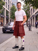 A man standing on a city sidewalk wearing a white t-shirt, a reddish-brown leather kilt with utility pockets, white knee-high socks, and black leather shoes. A black Mercedes-Benz car is parked on the street behind him, with trees and storefronts in the background.