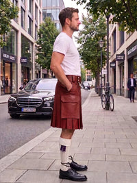 A man standing on a city sidewalk wearing a brown leather kilt, white t-shirt, white socks, and black leather shoes, with a street scene in the background.