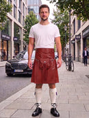 A man stands on a city sidewalk wearing a brown leather kilt with a front pouch, paired with a white t-shirt, white knee-high socks, and black shoes. The background shows a street scene with a black Mercedes-Benz car, storefronts, and trees.