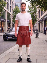 A man stands on a city sidewalk wearing a brown leather kilt with a front pouch, paired with a white t-shirt, white knee-high socks, and black shoes. The background shows a street scene with a black Mercedes-Benz car, storefronts, and trees.