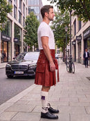 A man standing on a city sidewalk wearing a brown leather kilt, white t-shirt, white socks, and black leather shoes, with a street scene in the background.