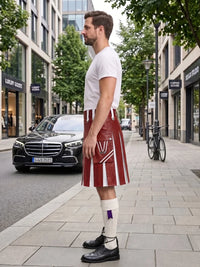 A man standing on a city sidewalk wearing a red leather kilt with white stripes, a white t-shirt, white knee-high socks, and black shoes. Buildings, trees, a parked black car, and a bicycle are visible in the background.