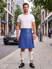 A man stands on a city sidewalk wearing a white crew-neck t-shirt and a blue pleated leather kilt with decorative metal studs. He is also wearing white knee-high socks and black leather shoes.