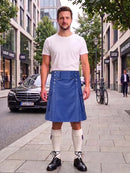 A man stands on a city sidewalk wearing a white crew-neck t-shirt and a blue pleated kilt with silver buttons, paired with white knee-high socks and black leather shoes. A black Mercedes-Benz is parked in the background on a city street.