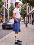 A man standing on a city sidewalk wearing a blue pleated kilt, white t-shirt, white socks, and black leather shoes, with buildings, trees, and parked cars in the background.