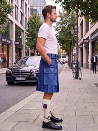A man standing on a city sidewalk wearing a blue pleated kilt, white t-shirt, white socks, and black leather shoes, with buildings, trees, and parked cars in the background.