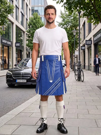 A man stands on a city sidewalk wearing a white crew-neck t-shirt and a blue leather kilt with white geometric piping details, paired with white knee-high socks and black leather shoes.