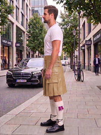A man standing on a city sidewalk wearing a beige kilt, white t-shirt, white knee-high socks, and black shoes, with a street scene and parked cars in the background.